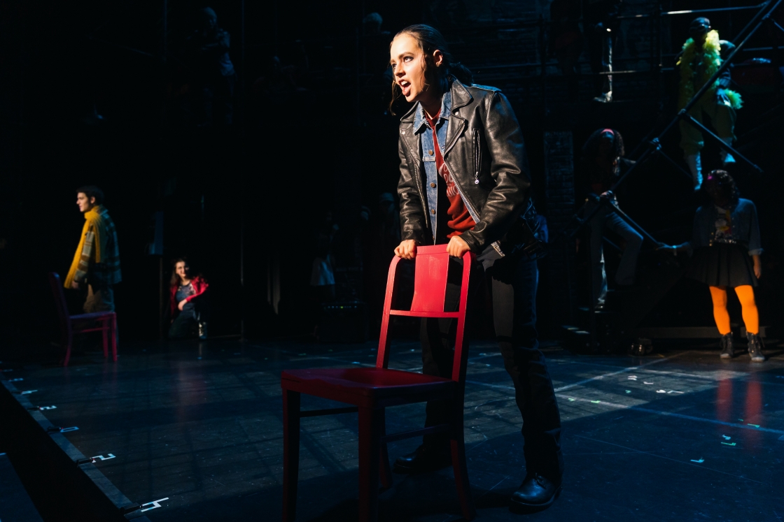 Actor in leather jacket singing while gripping the back of a red chair