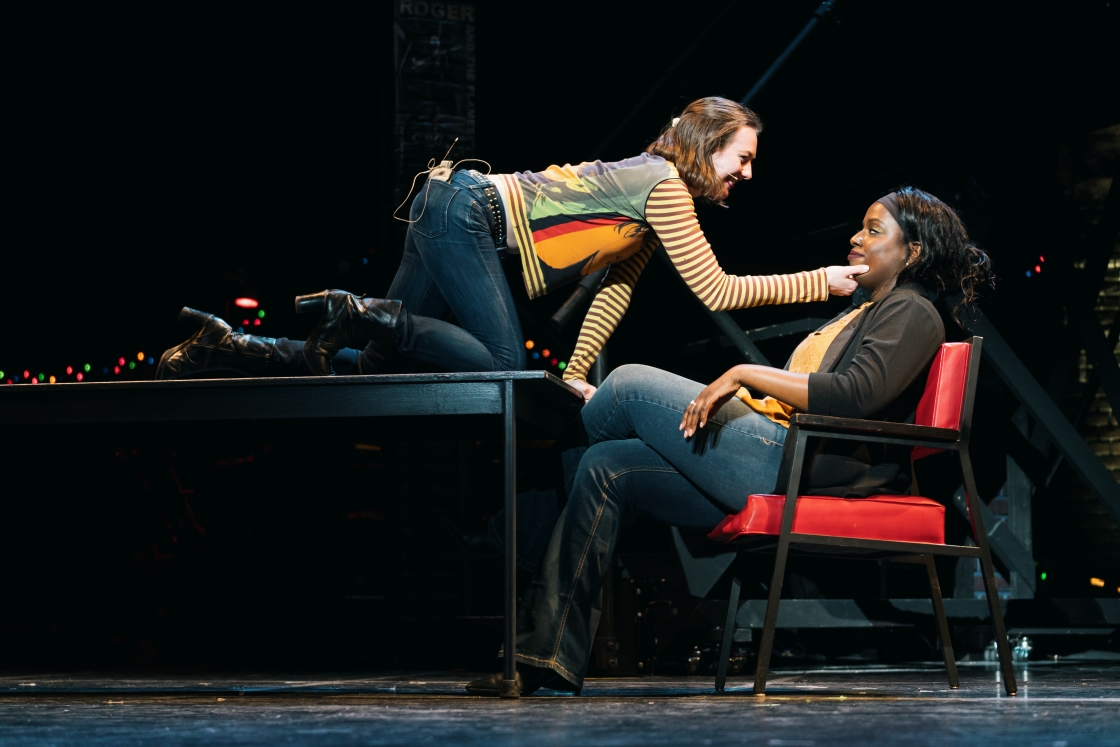 An actor kneeling on a table over a seated actor in Rent.