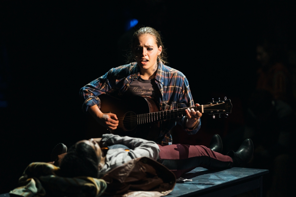 Actor in Rent singing and playing the guitar while seated next to a prone actor