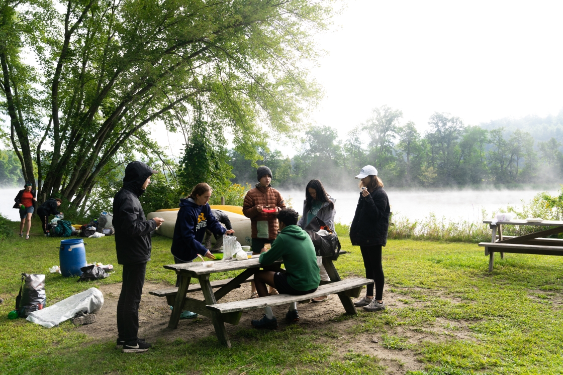 Students sit at a picnic table by the river with morning mist