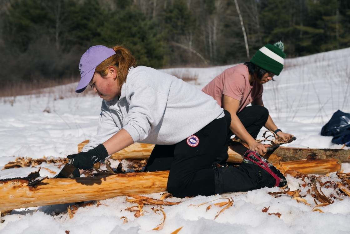 Two students kneel in the snow to scrape bark off hemlock logs