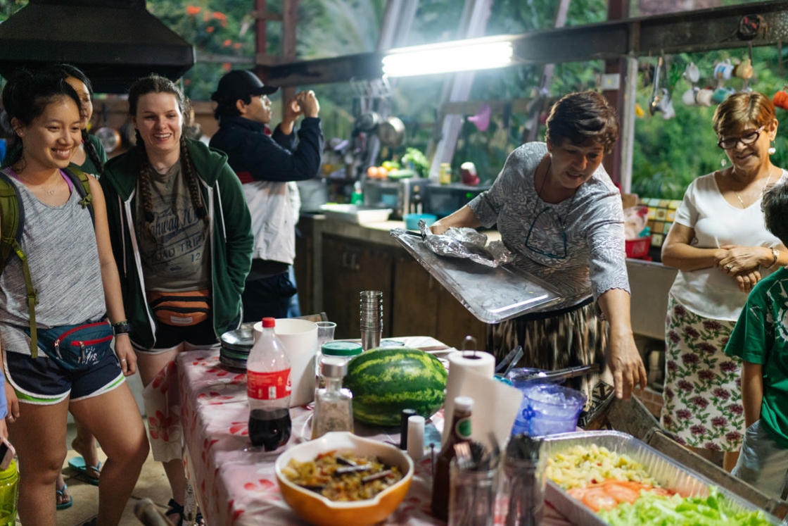 Two students look on as a women points out food that she has prepared for them in an outdoor kitchen in Puerto Rico