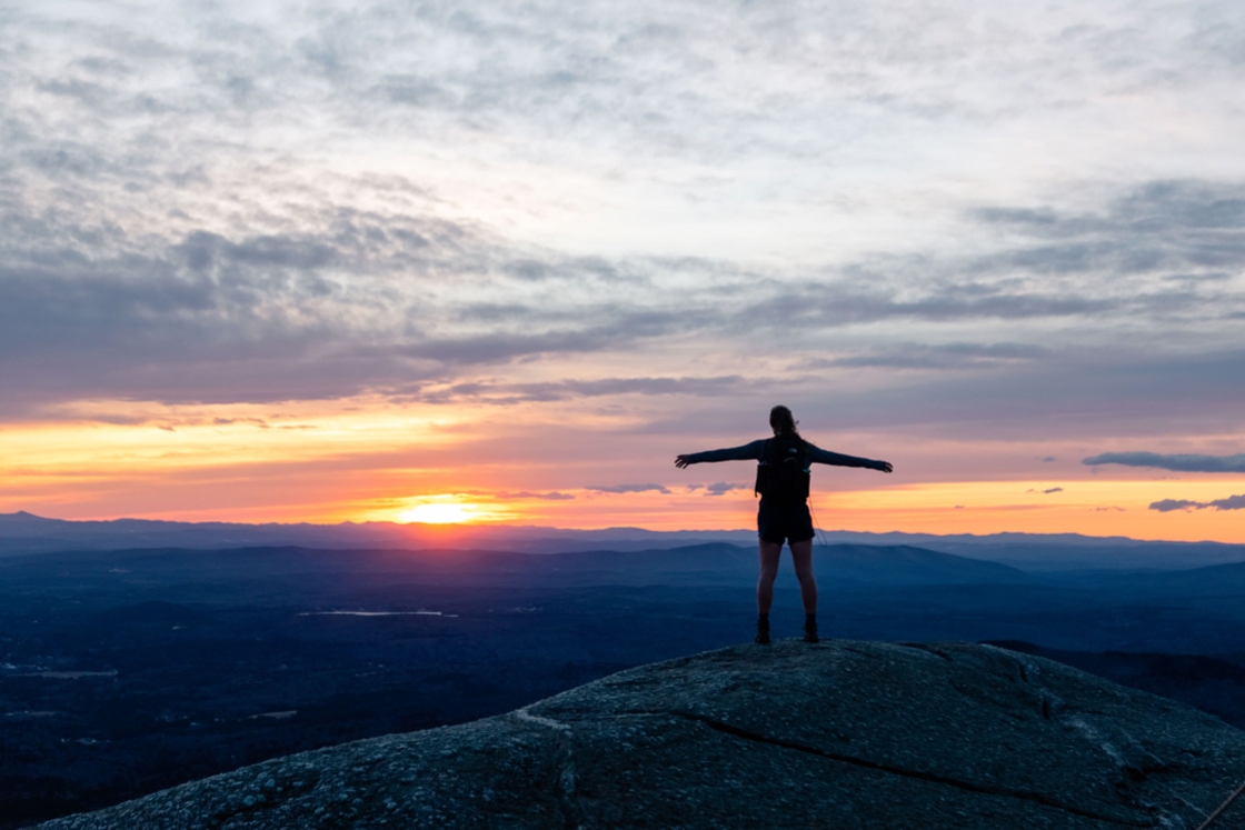 A hiker with arms outstretched watching the sunrise on the top of Mount Cardigan