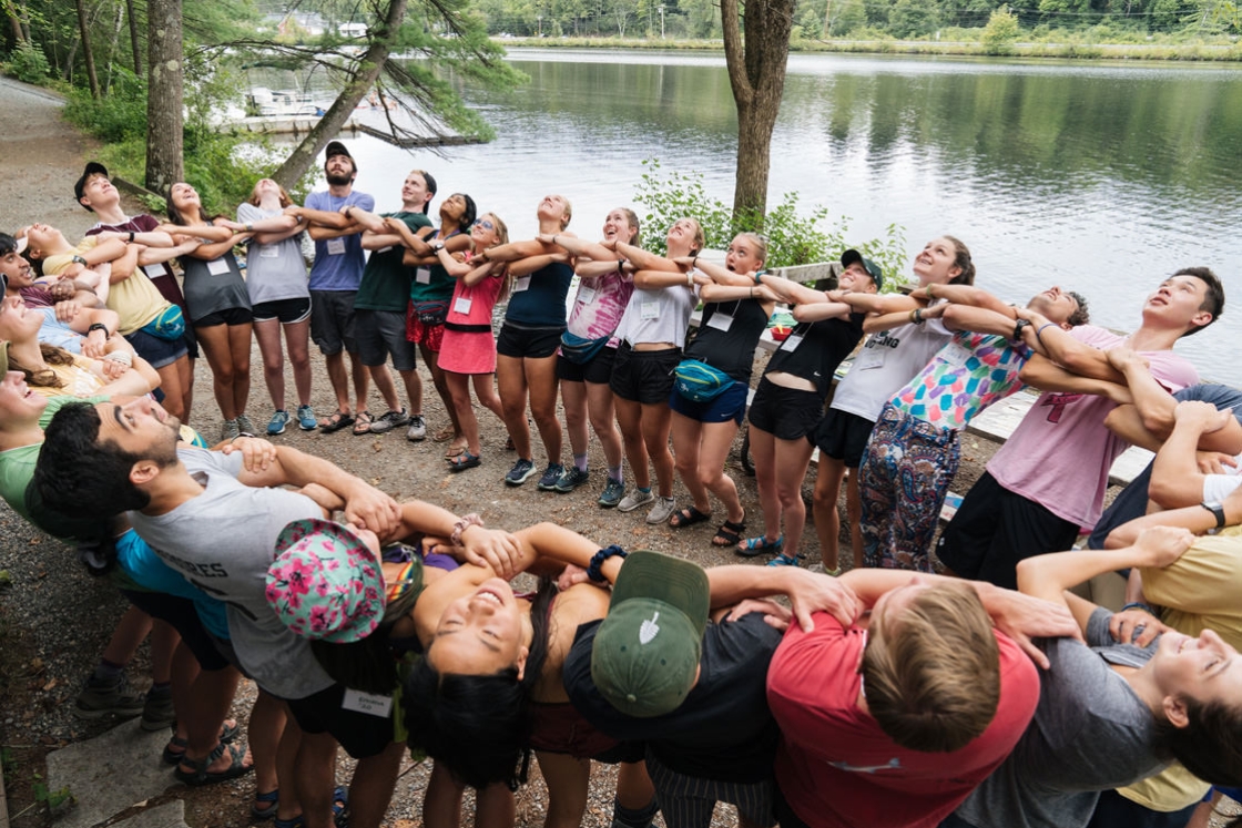 A group from the VOX croo leads first-years in team building exercise. Everyone stands in one circle, facing inward, with arms linked. They lean back and are responding to a call. They are standing next to the Connecticut River.