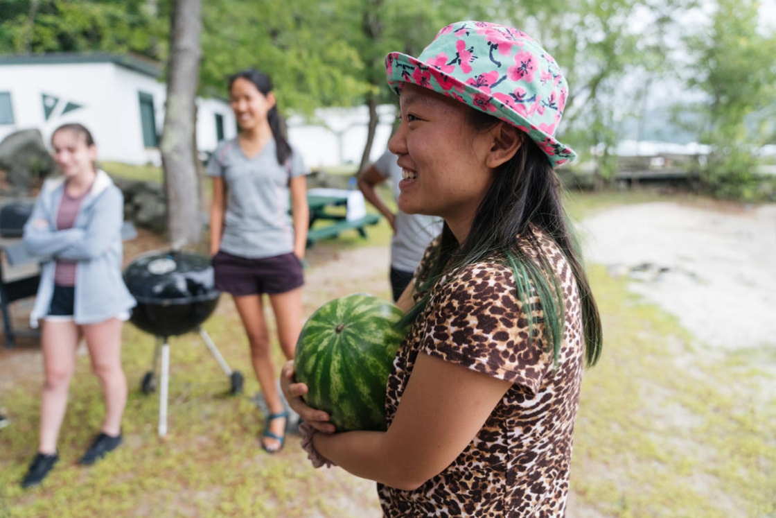 Emma Demers '20 holds a watermelon in her arms. She is smiling and is wearing a bright blue and pink hat.