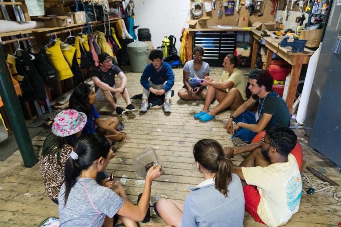 Members of Vox Croo sit in a boathouse in a circle on the ground and share a batch of brownies.