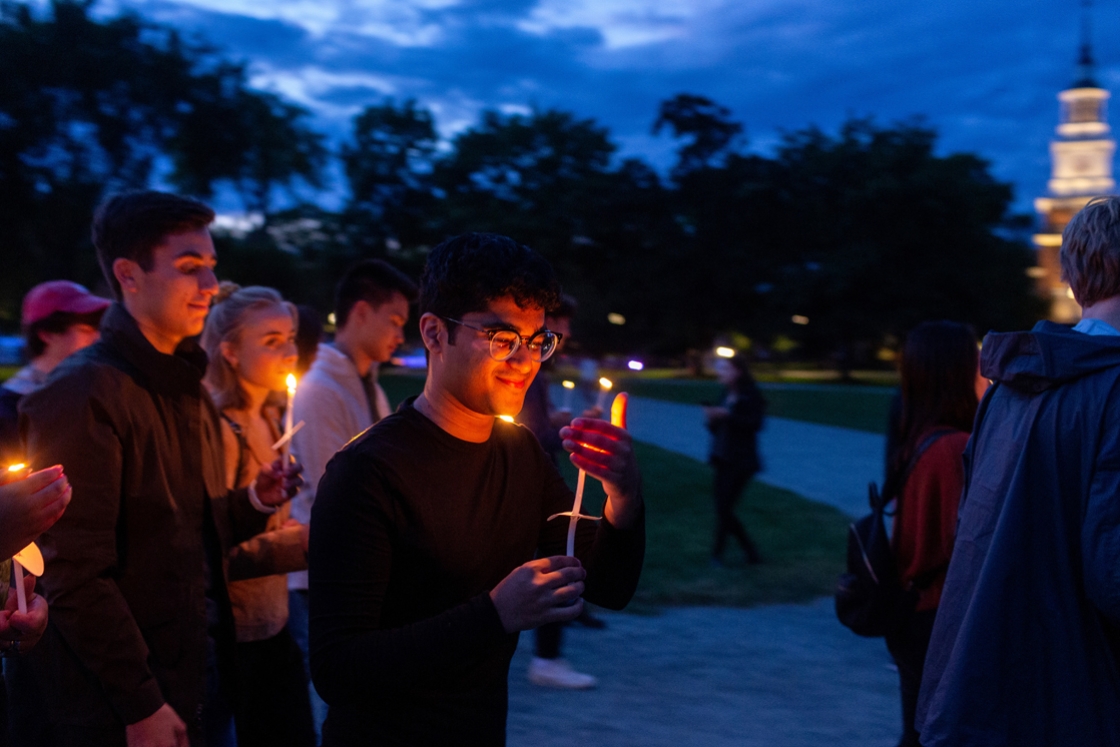 Taken at dusk, a first year holds a lit candle in front of them. Their face is lit up and in the background you can see Baker Library's lights. The student is a par