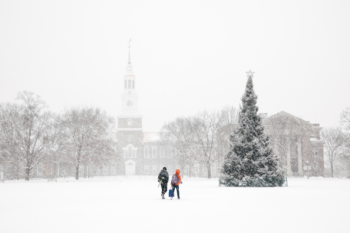 Two students walking in the snow past the Christmas tree on the Green