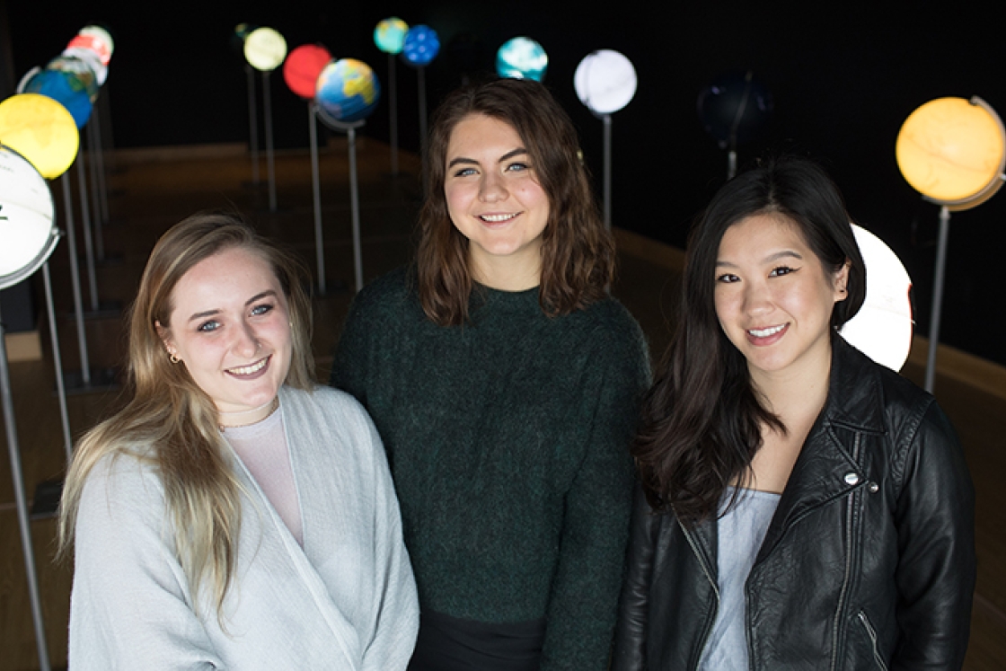 three female students standing together among an art installation of lit up globes