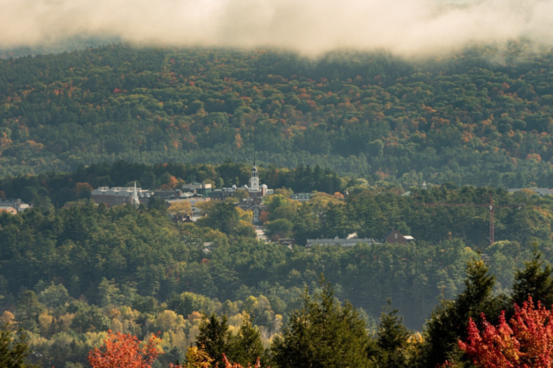 Aerial view of campus and Baker Library's tower from afar, surrounded by trees with turning leaves in fall