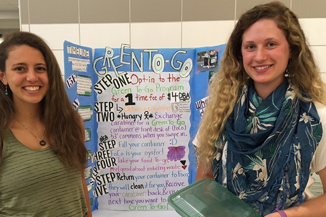 two female students standing in front of a poster, holding a green to-go box