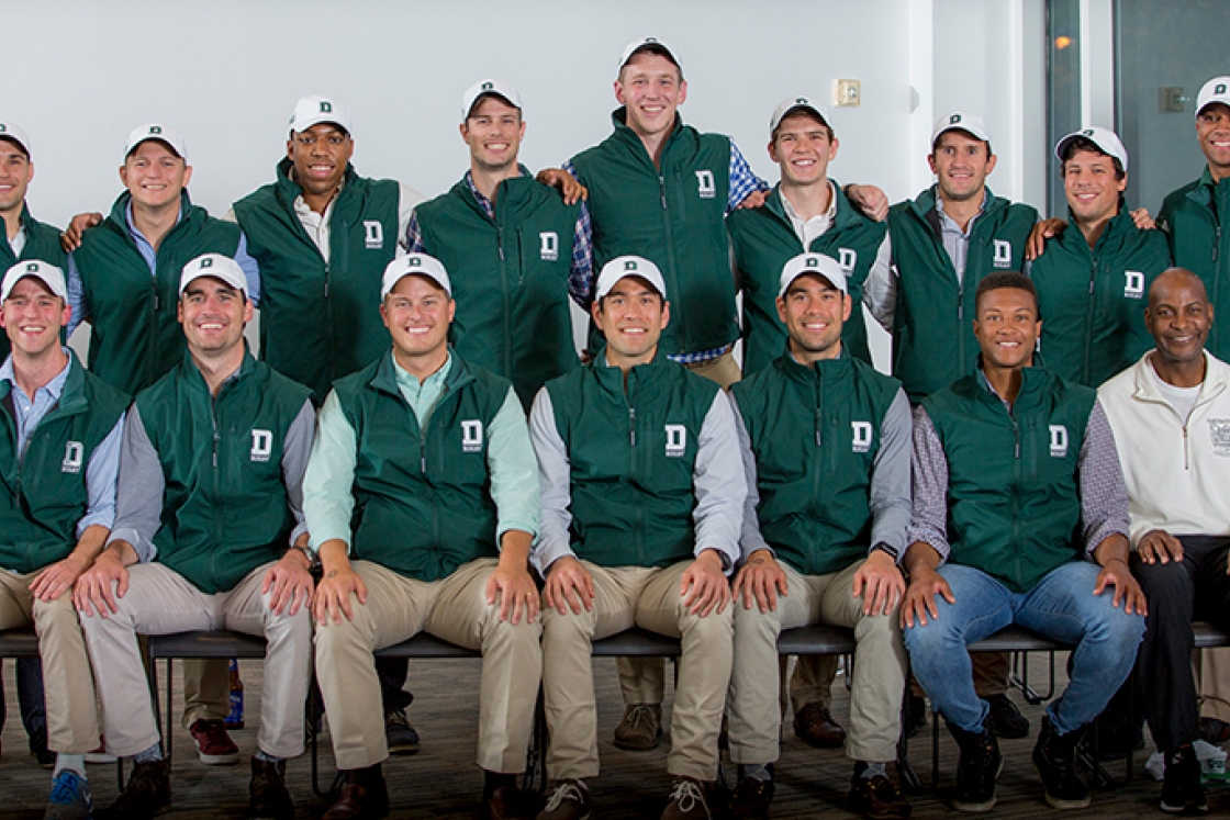 a group of male students, standing in two rows, wearing green vests with Dartmouth logos on them