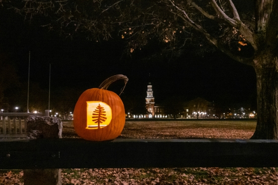 A pumpkin with a d carved in it sits on the sernior fence on the Green. It's nighttime and Baker-Berry Library can be seen in the distance.