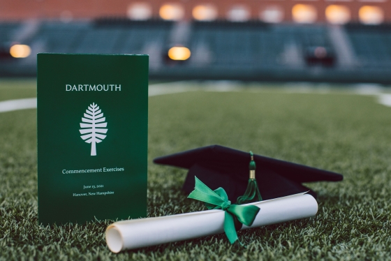 Commencement program, diploma rolled up with a green ribbon, and a mortarboard on the turf of Memorial Field