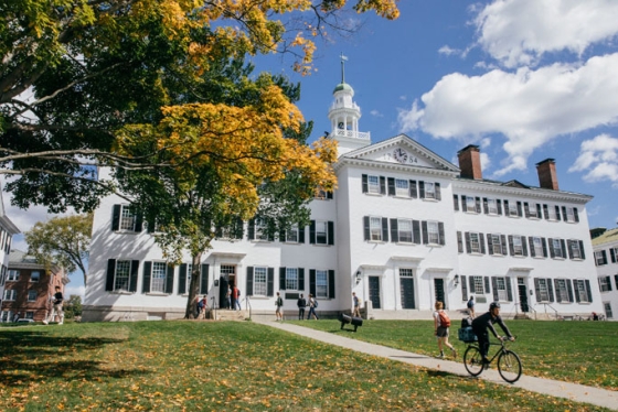 Dartmouth Hall during the fall. Students are walking to and from the hall and there is one riding his bike away from the hall. There is a tree with bright golden leaves.