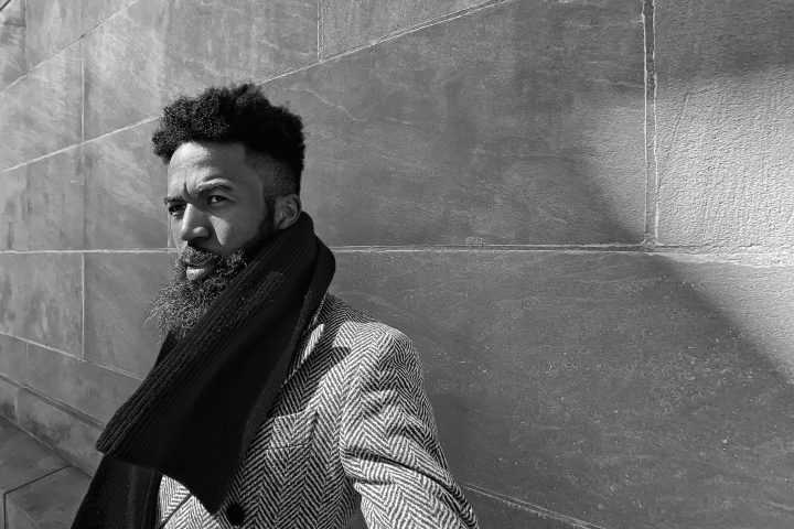 Black and white portrait of Joshua Bennett wearing a scarf and leaning against a slate wall