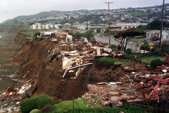Destroyed houses and debris at the edge of a cliff overlooking the ocean.