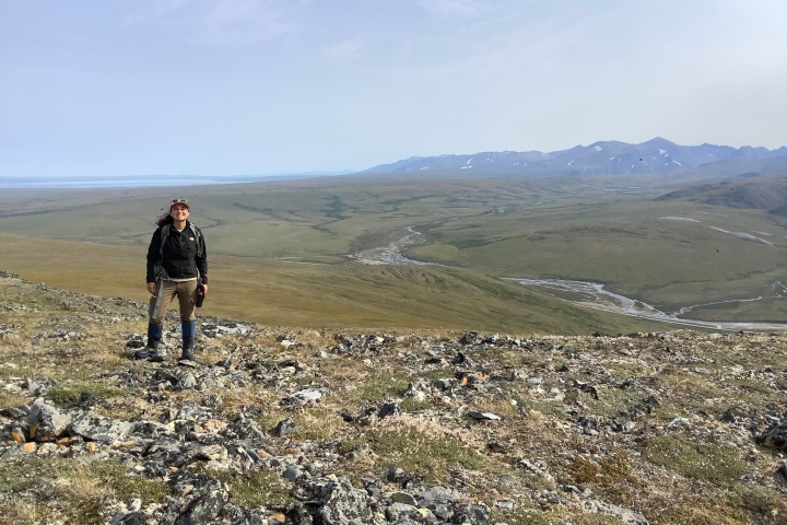 Woman in outdoor gear on rocky hilltop with mountains and wide river valley in background.