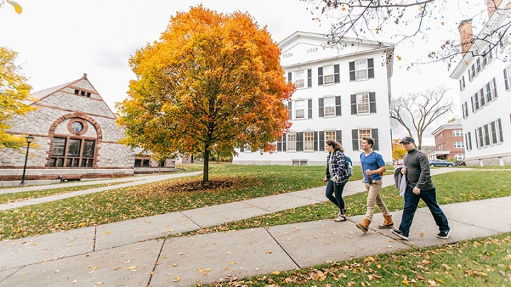 three students walking down the sidewalk near Rollins Chapel, a tree with yellow and orange leaves behind them