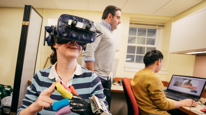 A student wears special gloves and a virtual reality headset while a student and professor watches her brainwaves on a computer monitor.
