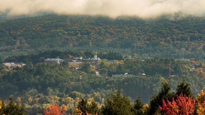 Aerial view of campus and Baker Library's tower from afar, surrounded by trees with turning leaves in fall