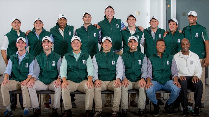 a group of male students, standing in two rows, wearing green vests with Dartmouth logos on them