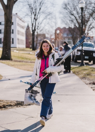 A student carries skis and boots across campus
